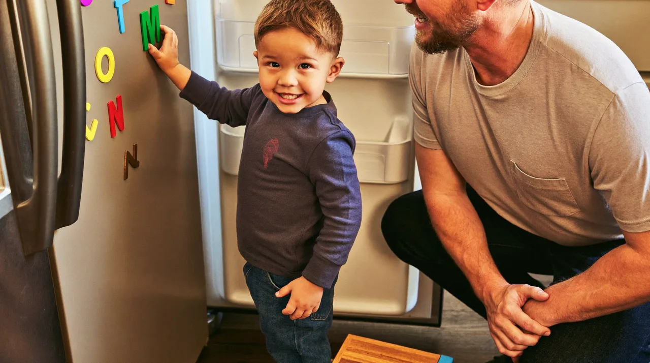 A young child presses a magnetic letter onto a fridge door while an adult watches and smiles.