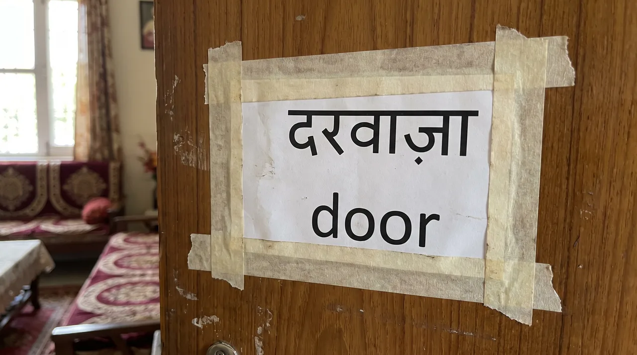 A small printed label taped to a household door showing the word 'door' written in Devanagari on the top line and in English on the bottom line.